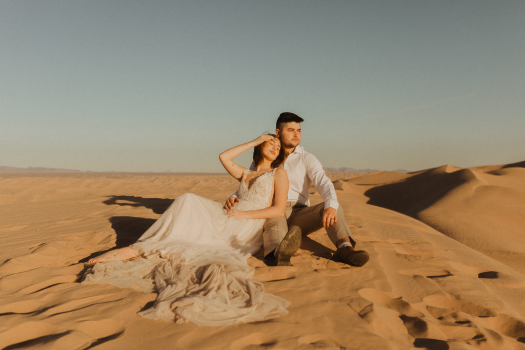 couple in wedding attire sitting on a sand dune